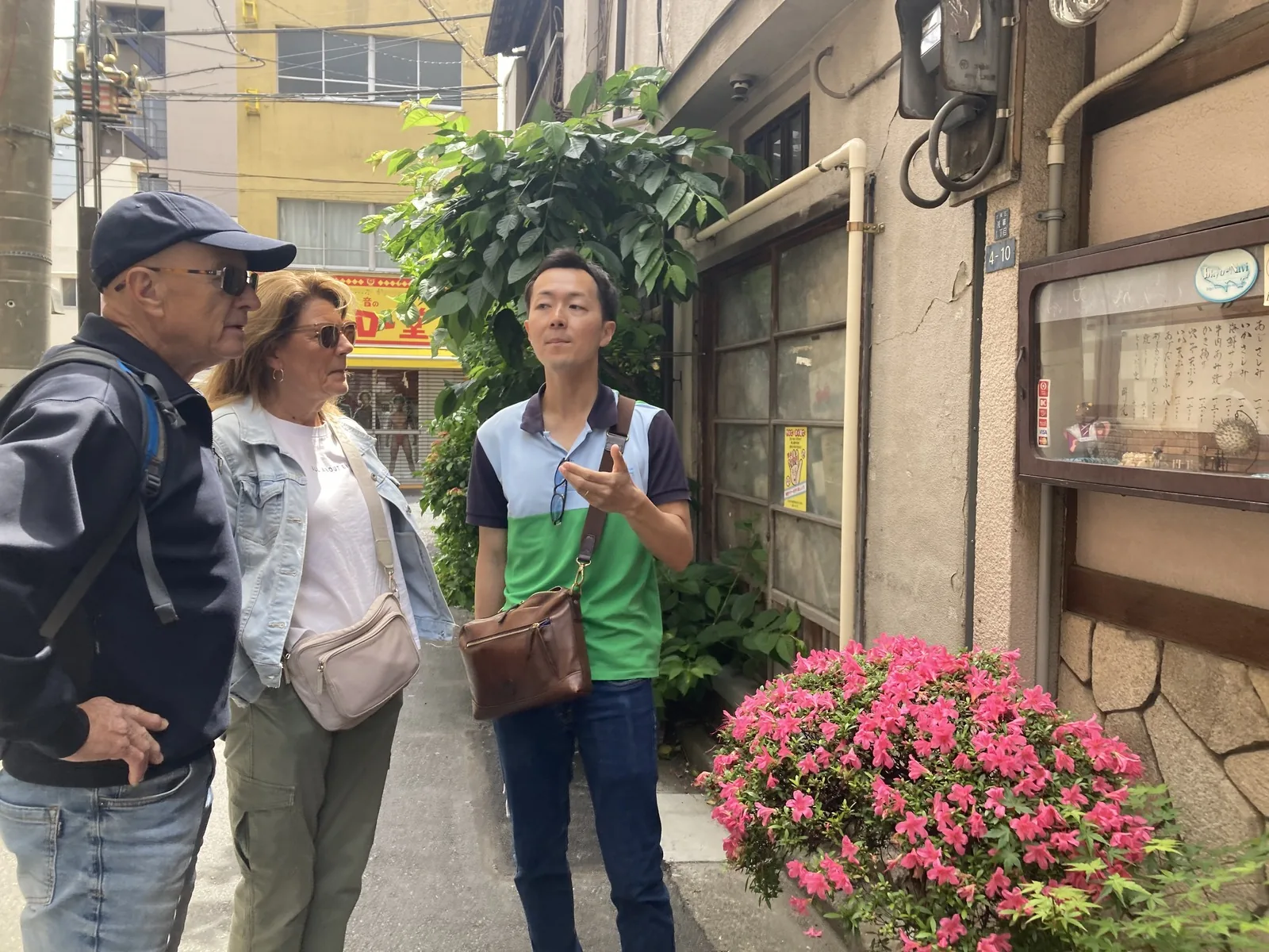 Manabu guiando a los visitantes por una calle trasera residencial tranquila en Asakusa con arbustos en flor